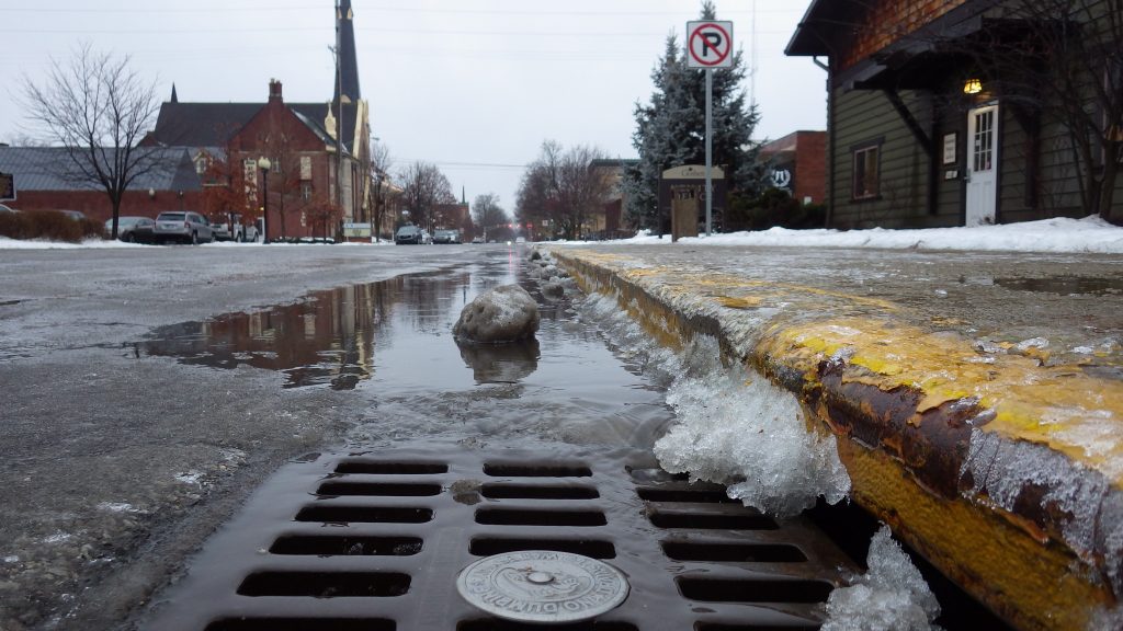 Up close look of a storm drain, snow and pooled water in the background